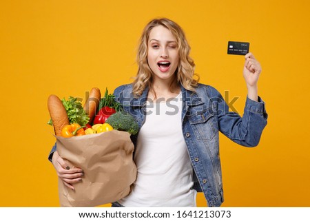 Blinking young girl in denim clothes isolated on orange background. Delivery service from shop or restaurant concept Hold brown craft paper bag for takeaway mock up with food product credit bank card