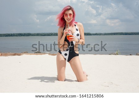 Young cheerful woman in swimsuit, toasting with cocktail, drinking fresh beverage on the beach, by sea. Vacation concept.