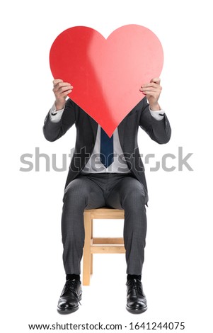  elegant businessman hiding behind big red heart, sitting isolated on white background, full body