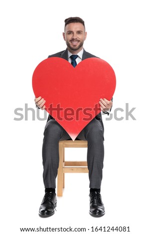 happy young businessman holding big red heart, sitting isolated on white background, full body
