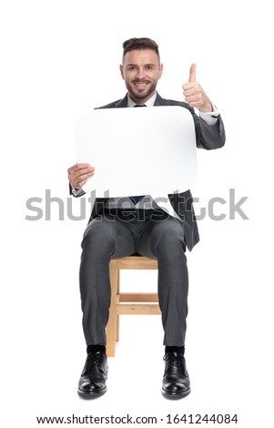 happy young businessman holding speech bubble and making thumbs up sign, sitting isolated on white background, full body