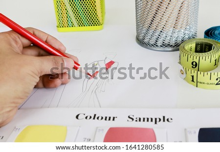 Hand holding red color pencil is sketching fashion design  with color  pencils, fabric color sample and yellow tape measure on the table, focus at pencil tip.