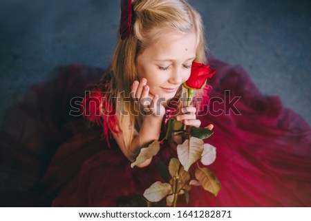 Llittle girl with blond hair holds a gorgeous red rose. Red luxurious dress. Valentine's Day.