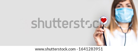young woman in a medical mask holds a stethoscope head with a heart on a light background. Added pulse and heart icon. Banner. Doctor concept, high level medical, vaccination, visit to the doctor