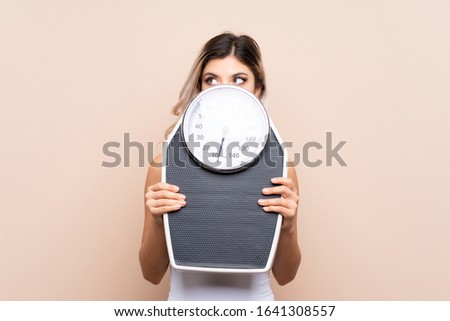 Teenager girl with weighing machine over isolated background with weighing machine and hiding behind it