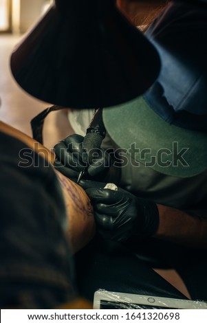 Close up of professional tattooer artist doing picture on hand of man by machine black ink from a jar. 