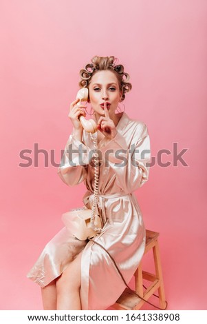 Housewife in pink robe gossiping on phone and puts her finger to her mouth.