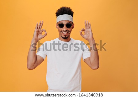 Young man in headband and stylish glasses shows sign ok. Portrait of brunette man in sunglasses and white tee smiling and posing on orange background