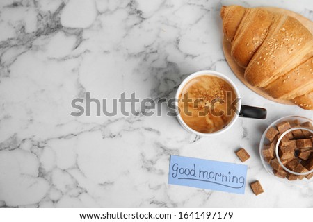 Delicious coffee, croissant and card with words GOOD MORNING on white marble table, flat lay. Space for text