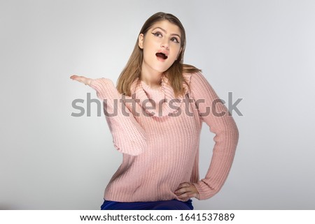 Close up portrait of a young Caucasian woman wearing a pink sweater on a grey background.  She is presenting something like and advertisement