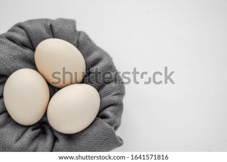 Top view of three unpainted beige eggs in the nest of the grey fabric on a white background.
