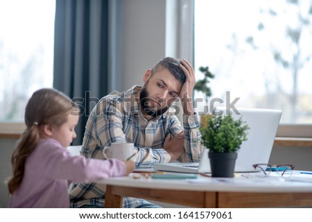 Daddys care. Tired young dad looking at the drawing little daughter, both sitting at the table at home.