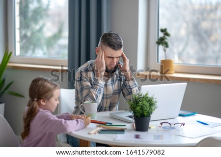 Tired dad. Tired sad dad, his hands at his temples, a little daughter with pencils, both sitting at a white table.