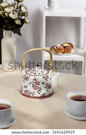 Subject shot of a white teapot with pink roses print and a straw handle. A vase with white flowers and tea cups are on the beige table-cloth against the white whatnot with tea dish and plain rolls. 