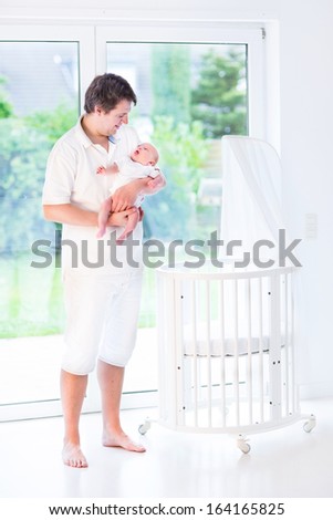 Young smiling father holding his newborn baby standing next to a white round crib with canopy at a big window