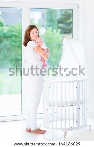Young mother holding her newborn baby standing at a white crib next to a big window