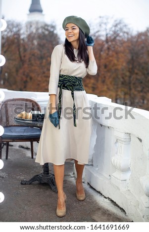 Beautiful young woman standing on a classic balcony, wearing light beige spring trench coat and beret hat, looking at camera and smiling