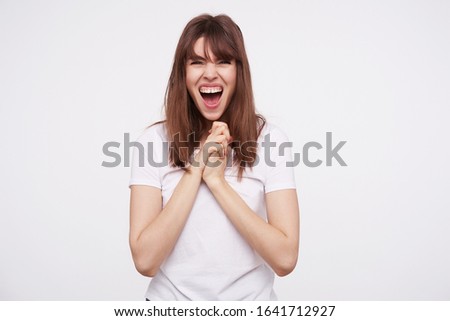 Excited young pretty dark haired female keeping raised hand under her chin and screaming with wide mouth opened, posing over white background in casual clothes Royalty-Free Stock Photo #1641712927