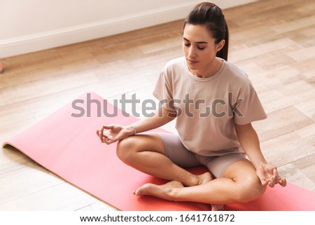 Image of a young beautiful brunette fitness woman sit on rug indoors at home meditate.