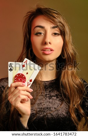 Brunette lady in black dress showing two red chips and aces, posing against colorful studio background. Gambling, poker, casino. Close-up.