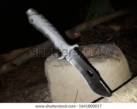 Black survival knife stuck in a tree in front of a clay stove in the forest 