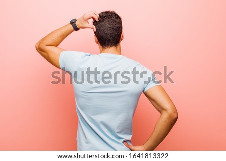 young arabian man feeling clueless and confused, thinking a solution, with hand on hip and other on head, rear view against pink wall
