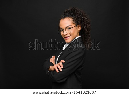 young black woman smiling to camera with crossed arms and a happy, confident, satisfied expression, lateral view against black wall