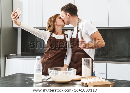 Happy young couple wearing aprons standing at the kitchen desk, taking a selfie while making dough