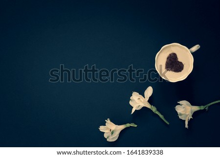 Minimalistic flat lay. Three flowers and coffee with cream and cinnamon in the form of a heart in a mug on a dark blue background. White with yellow petals. Vignetting. Copy space on the left.