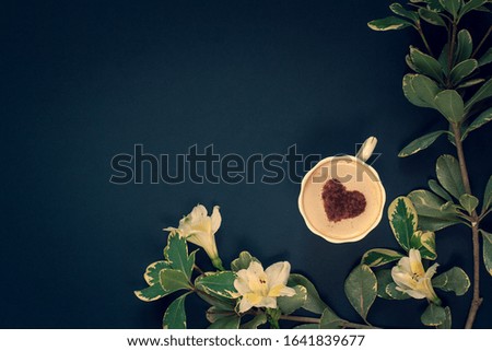 Minimalistic flat lay. Coffee mug with cream and cinnamon in the form of a heart with flowers and a branch on a dark blue background. White with yellow petals. Vignetting. Copy space on top.