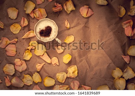 Coffee mug with cream and cinnamon in the form of a heart with pink flower petals on crumpled kraft paper. Flat lay. Copy space. Vignetting.