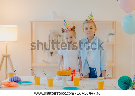 Glad boy and his small ginger sister dressed in festive clothes, party hats, celebrate birthday together, surrounded with cake, present and cups of drink, have good mood during special occasion