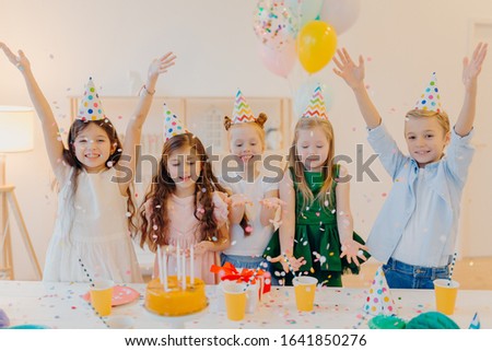 Horizontal shot of happy positive children catch confetti, celebrate birthday together, raise arms, have good mood, play together, stand near festive table with present box, cake, party hats