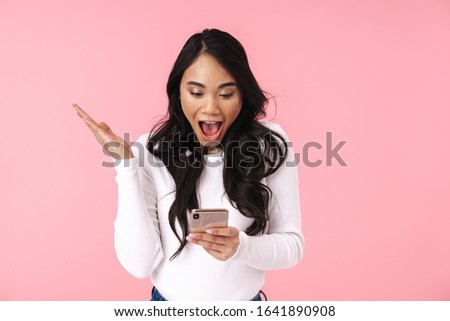 Image of young brunette asian woman with long hair wondering and holding cellphone isolated over pink background