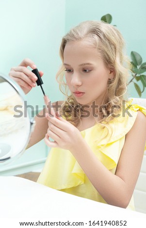 young blonde girl sits at a table and paints her lips
