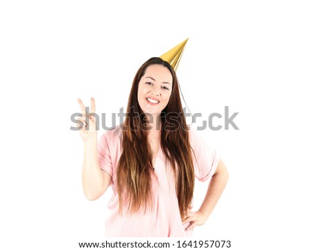 Cute young woman smiling and doing a two finger sign with her hand while wearing a gold party hat against a white background