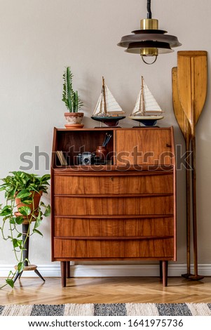 Stylish and vintage interior design of living room with wooden retro commode,  plants, ships, paddle, lamp and elegant personal accessories. Template. Beige wall. Vintage concept of home decor.