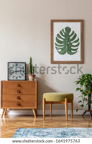 Vintage interior design of living room with design retro pouf and commode, plants, cacti, black clock and brown  mock up poster frame on the beige wall. Stylish home decor. Template. 