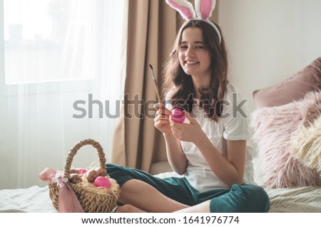 Teenager girl with Easter ears and paints easter eggs with a brush  on a bed in a living room. Emotions. Easter concept