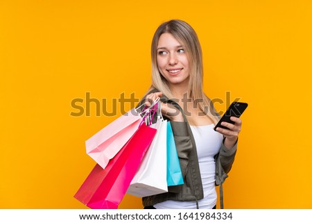 Young blonde woman over isolated yellow background holding shopping bags and a mobile phone