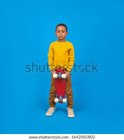 Cute African-American teenager in casual clothes, yellow sweater and brown pants, standing and holding red skateboard and smiling against blue background. Concept of activity and happy childhood.