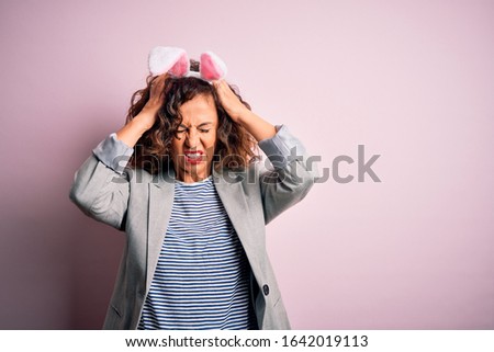 Middle age beautiful woman wearing bunny ears standing over isolated pink background suffering from headache desperate and stressed because pain and migraine. Hands on head.