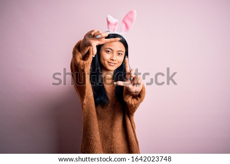 Young beautiful chinese woman wearing bunny ears standing over isolated pink background smiling making frame with hands and fingers with happy face. Creativity and photography concept.