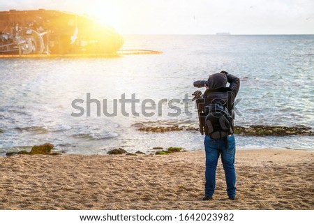 Photographer traveler with a camera and a backpack on the beach in spring when it is still cold, takes pictures sea.