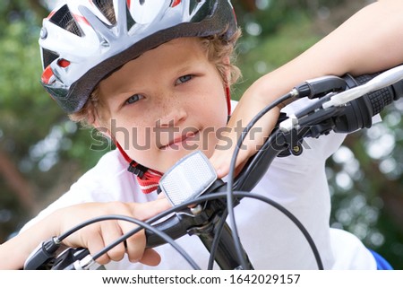 Close-up of a teenager boy in protective helmet sitting on his bicycle and keeps his head on his hands resting on the handlebars of the bike in park on summer day.