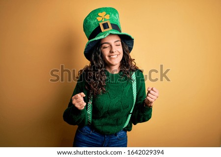 Beautiful curly hair woman wearing green hat with clover celebrating saint patricks day excited for success with arms raised and eyes closed celebrating victory smiling. Winner concept.