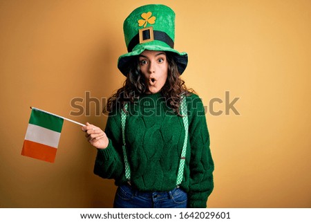 Beautiful patriotic curly hair woman wearing hat holding irish flag celebrating saint patricks day scared in shock with a surprise face, afraid and excited with fear expression