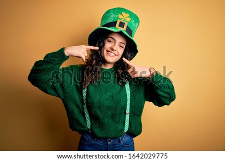 Beautiful curly hair woman wearing green hat with clover celebrating saint patricks day smiling cheerful showing and pointing with fingers teeth and mouth. Dental health concept.