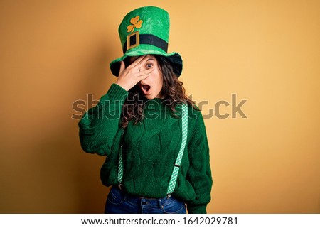 Beautiful curly hair woman wearing green hat with clover celebrating saint patricks day peeking in shock covering face and eyes with hand, looking through fingers with embarrassed expression.
