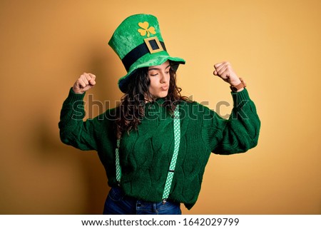 Beautiful curly hair woman wearing green hat with clover celebrating saint patricks day showing arms muscles smiling proud. Fitness concept.